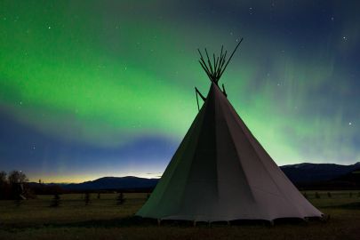 The northern lights dance as the sun rises behind a teepee in the Yukon.